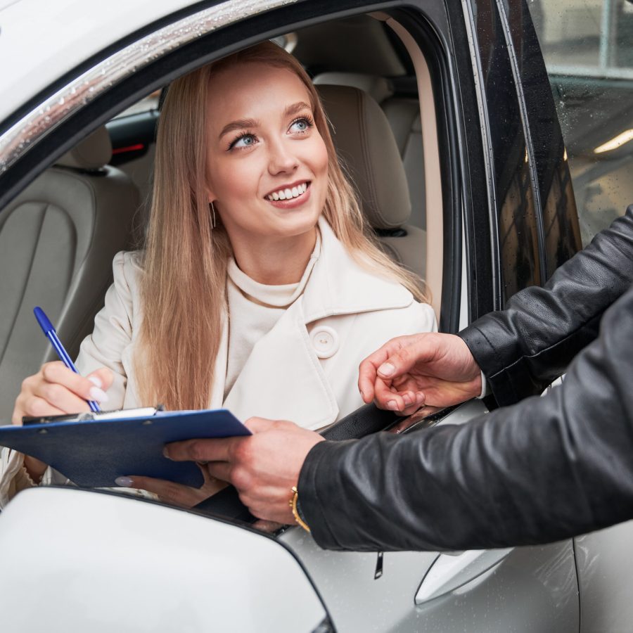 Close-up view of beautiful lady sitting inside white car, looking out of lowered window at man who showing her documents needed to sign. Business woman signing agreement in salon of her auto.